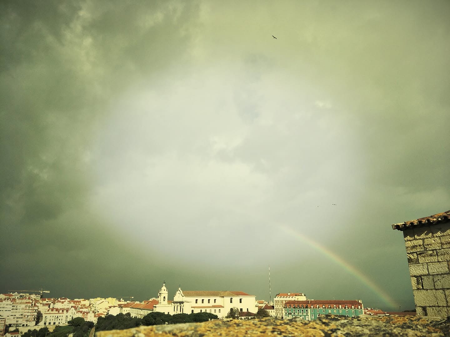 A rainbow above the rooftops of Lisbon beneath a green-gray sky.